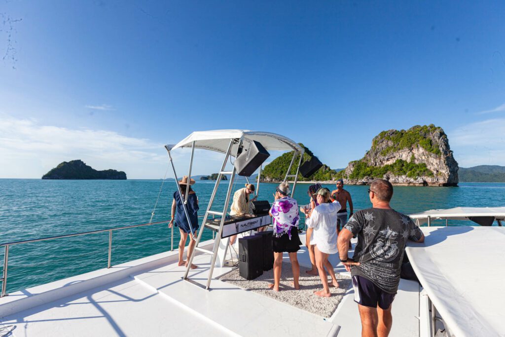 A DJ mixing while a crowd stands around him on board a party boat